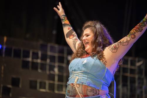 Aktorka w krótkim dżinsowym topie, z licznymi tatuażami unosi ręce ku górze, w geście zadowolenia.